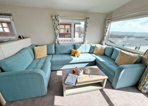 A modern living room with a teal sectional sofa, a wooden coffee table with books and a teapot, and large windows offering a view of the countryside. - Valley View 2 Bed Holiday Caravan Rame (St Ann&rsquo;s Chapel, Callington)
