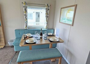 A dining area with a table set for two, a blue cushioned bench, a window with curtains, and a framed picture on the wall. - Valley View 2 Bed Holiday Caravan Rame (St Ann&rsquo;s Chapel, Callington)