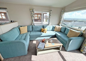 Ein modernes Wohnzimmer mit einem salbeengrünfarbenen Ecksofa, einem Couchtisch aus Holz mit Büchern und einer Teekanne sowie großen Fenstern mit Blick auf die Landschaft.