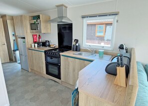 A compact kitchen with a stove, oven, microwave, and sink. There is a window with blinds, a door leading to another room, and a countertop with a knife block. - Valley View 2 Bed Holiday Caravan Rame (St Ann&rsquo;s Chapel, Callington)