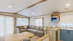 En kompakt opholdsstue med sofa, et lille bord, et tekøkken med mikroovn og et vindue med gardiner.