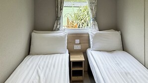 A small, single-room accommodation with two single beds, a wooden nightstand, and a window with curtains. - Luxury caravan with one double and one twin room. (Llanrhystud, Cardiganshire)