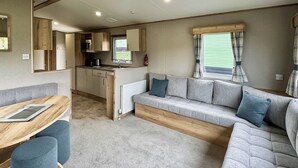 A caravan interior with a kitchenette, a sofa, a dining table, and a window with curtains. - Luxury caravan with one double and one twin room. (Llanrhystud, Cardiganshire)
