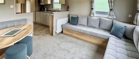 Interior de una caravana con kitchenette, sofá, mesa de comedor y ventana con cortinas.