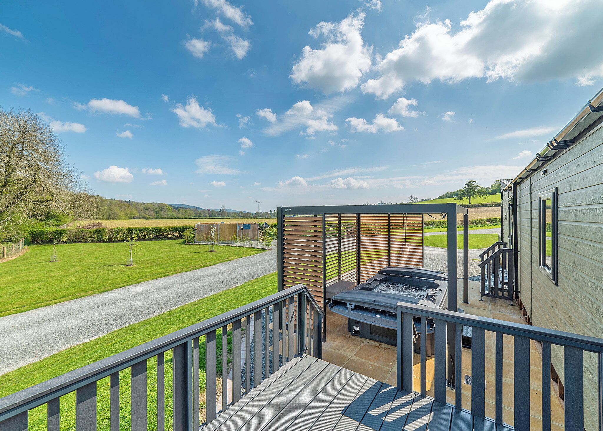 A patio area with a hot tub, a wooden deck, and a view of a green field and distant hills.
