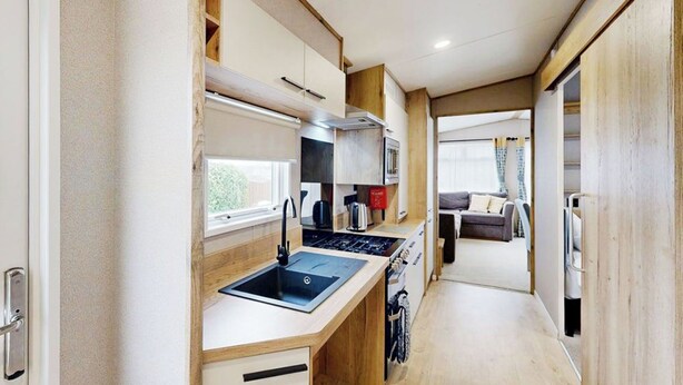 Una kitchenette compacta con pileta, estufa y alacenas, que da a una sala de estar con sofá y ventana.