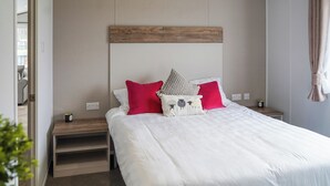 Un lit bien fait, avec une literie blanche et deux oreillers, une tête de lit en bois, une table de chevet avec une lampe et une prise de courant fixée au mur.