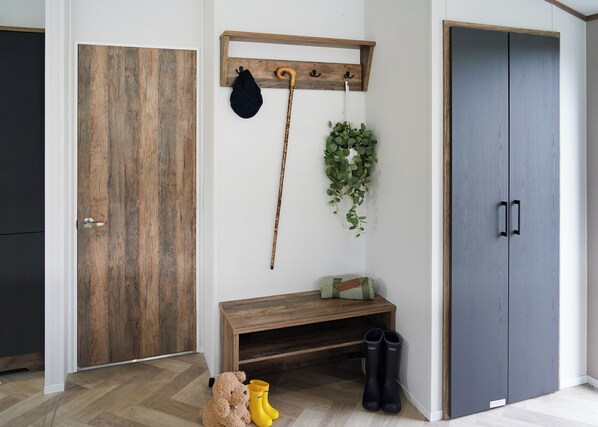 Una habitación con puerta de madera, un perchero con sombrero y bastón, un banco con botas y un armario gris.