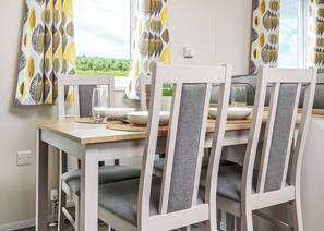 A dining area with a wooden table, white chairs with grey cushions, and patterned curtains. - Signature 2 Bed Caravan Wheelchair Accessible (Ormside, Appleby-in-Westmorland)
