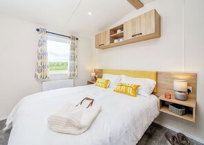 A bedroom with a bed, bedside tables, a headboard, a window with curtains, a radiator, and a wooden shelf above the bed. - Signature 2 Bed Caravan Wheelchair Accessible (Ormside, Appleby-in-Westmorland)