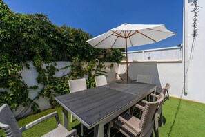 Eine Terrasse mit Tisch, Stühlen und einem Sonnenschirm, sowie eine weiße Wand mit Grünpflanzen.