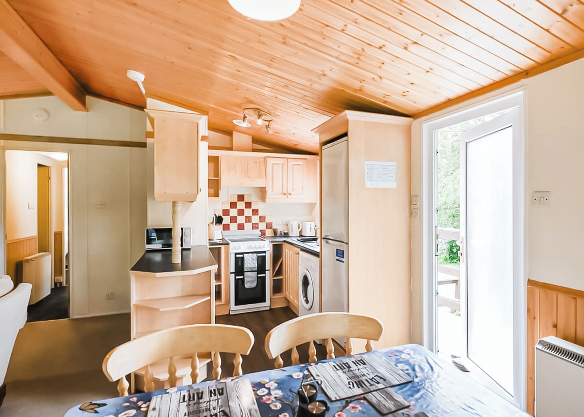 A compact kitchen with wooden cabinets, a stove, and a refrigerator. There is a dining table with chairs and a view of greenery outside.