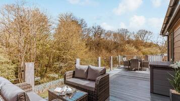 A deck with wicker furniture, a glass-top coffee table, and a view of trees and a building.