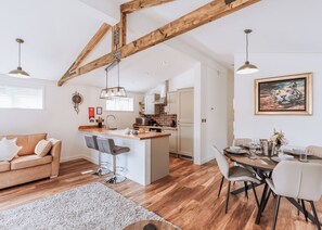 Une cuisine moderne avec des poutres en bois, un coin repas avec une table dressée pour deux personnes, et un espace de vie chaleureux avec un canapé et des tabourets de bar.