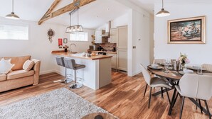 Cozinha moderna com vigas de madeira, área de refeições com mesa posta para dois e um espaço de estar acolhedor com sofá e banquetas.