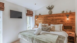 Ein Schlafzimmer mit Holz-Kopfteil, Flachbildfernseher, Nachttisch mit Lampe und einem Vorhangfenster.