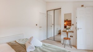 Um quarto com uma cama, um espelho, um banquinho e uma porta que dá para outro cômodo.
