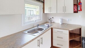 A kitchen with a built-in sink, white cabinets, a window, and a fire extinguisher. - Gold Plus 3 Sleeps 6 (Pet) (New Quay)