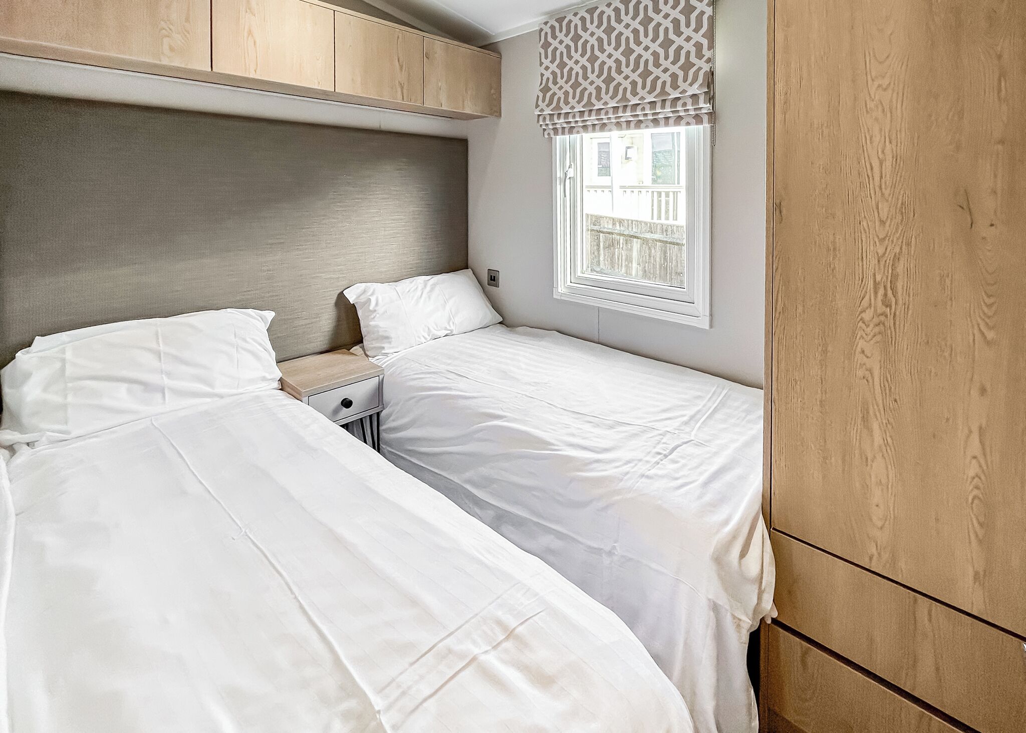 A bedroom with two beds, wooden headboard, bedside tables, a window with a patterned valance, and a view of the outside balcony.
