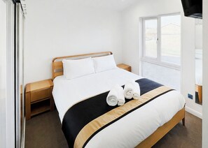 A hotel room with a bed, wooden headboard, white bedding, two rolled towels, a nightstand, a window with blinds, and a television on the wall. - Executive Lodge with Hot Tub (Monkton, Nr Ramsgate)