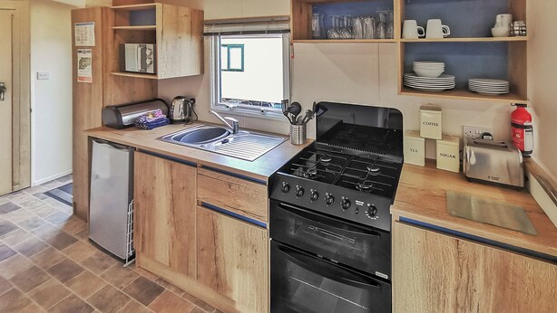 A compact kitchen with a built-in stove, sink, and a small refrigerator. - Gold Plus 3 Sleeps 6 (New Quay)