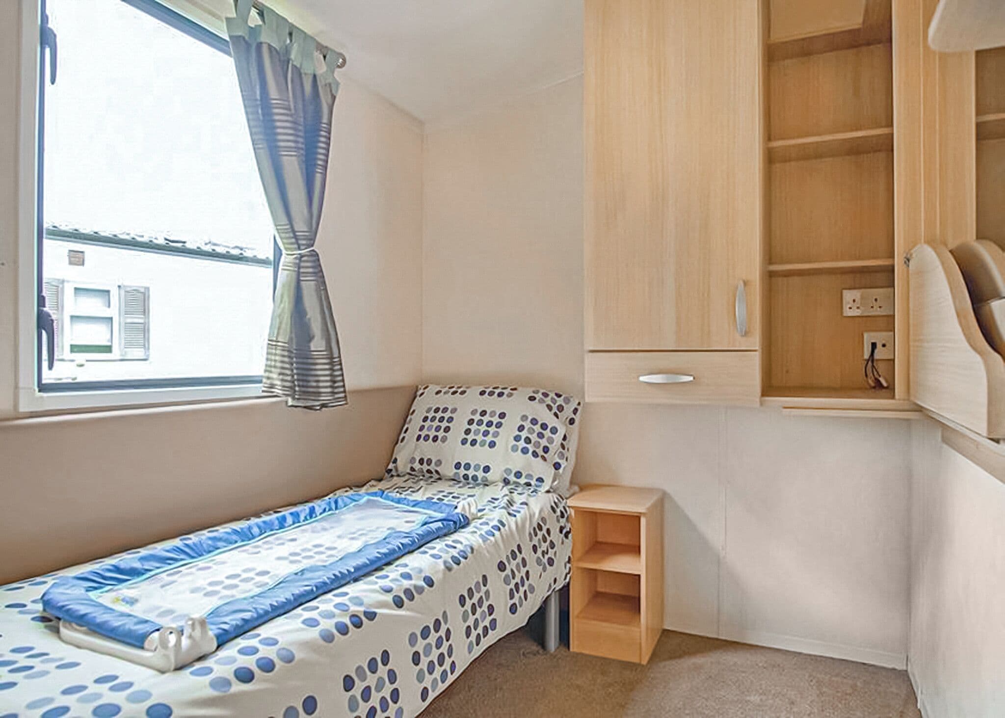 A small bedroom with a single bed, a wooden cabinet, a small shelf, and a window with curtains.