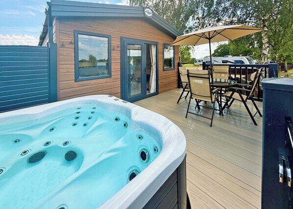 A hot tub on a wooden deck with a patio table and chairs, a covered umbrella, and a small building with windows.