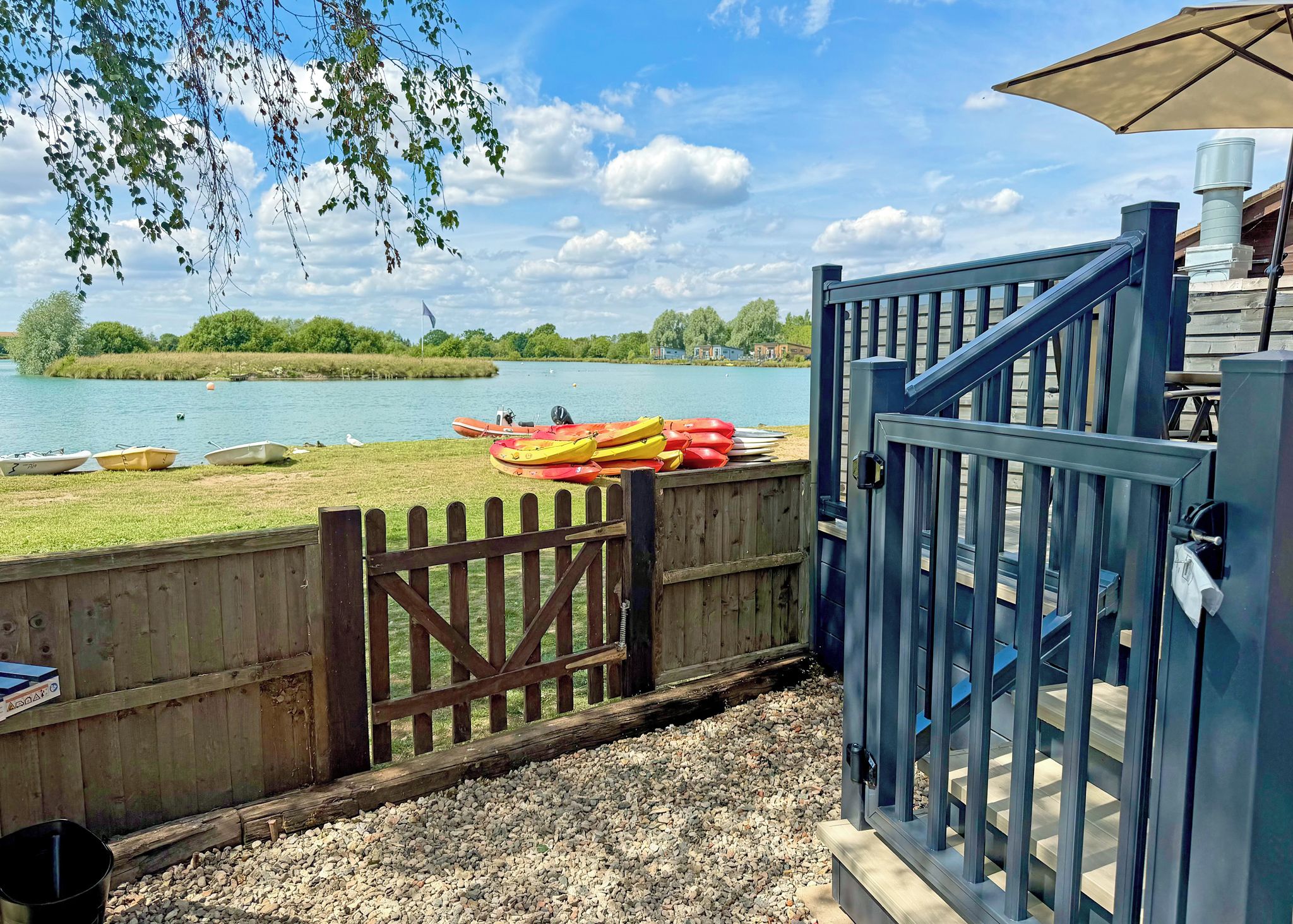 A wooden dock with kayaks and a boat, a wooden fence, and a blue gate leading to a building with a staircase.