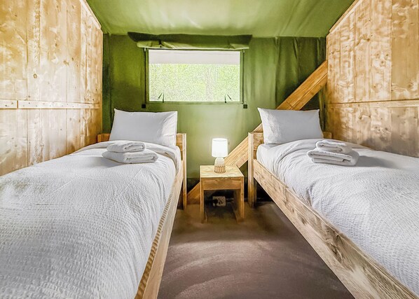 Two wooden bunk beds with white bedding, a nightstand with a lamp, and a window with a view of trees.