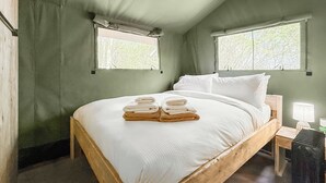 A wooden bed with white linens and towels, two windows with curtains, a bedside table with a lamp, and a radiator.