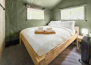 Ein hölzernes Bett mit weißen Leinentüchern und Handtüchern, zwei Fenster mit Vorhängen, ein Nachttisch mit Lampe und ein Heizkörper.