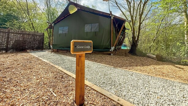 A wooden sign labeled 'Goose Lodge' in front of a green building with a slanted roof, surrounded by trees and gravel paths.