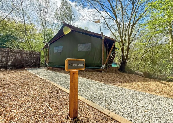 A wooden sign labeled 'Goose Lodge' in front of a green building with a slanted roof, surrounded by trees and gravel paths.