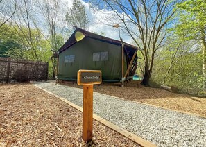 Ein hölzernes Schild mit der Aufschrift 'Goose Lodge' vor einem grünen Gebäude mit einem schrägen Dach, umgeben von Bäumen und Kieswegen.