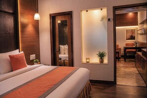 Habitació d'hotel amb un llit, una taula de nit, una làmpada de paret, una porta que dona a un bany i una petita finestra amb una planta.