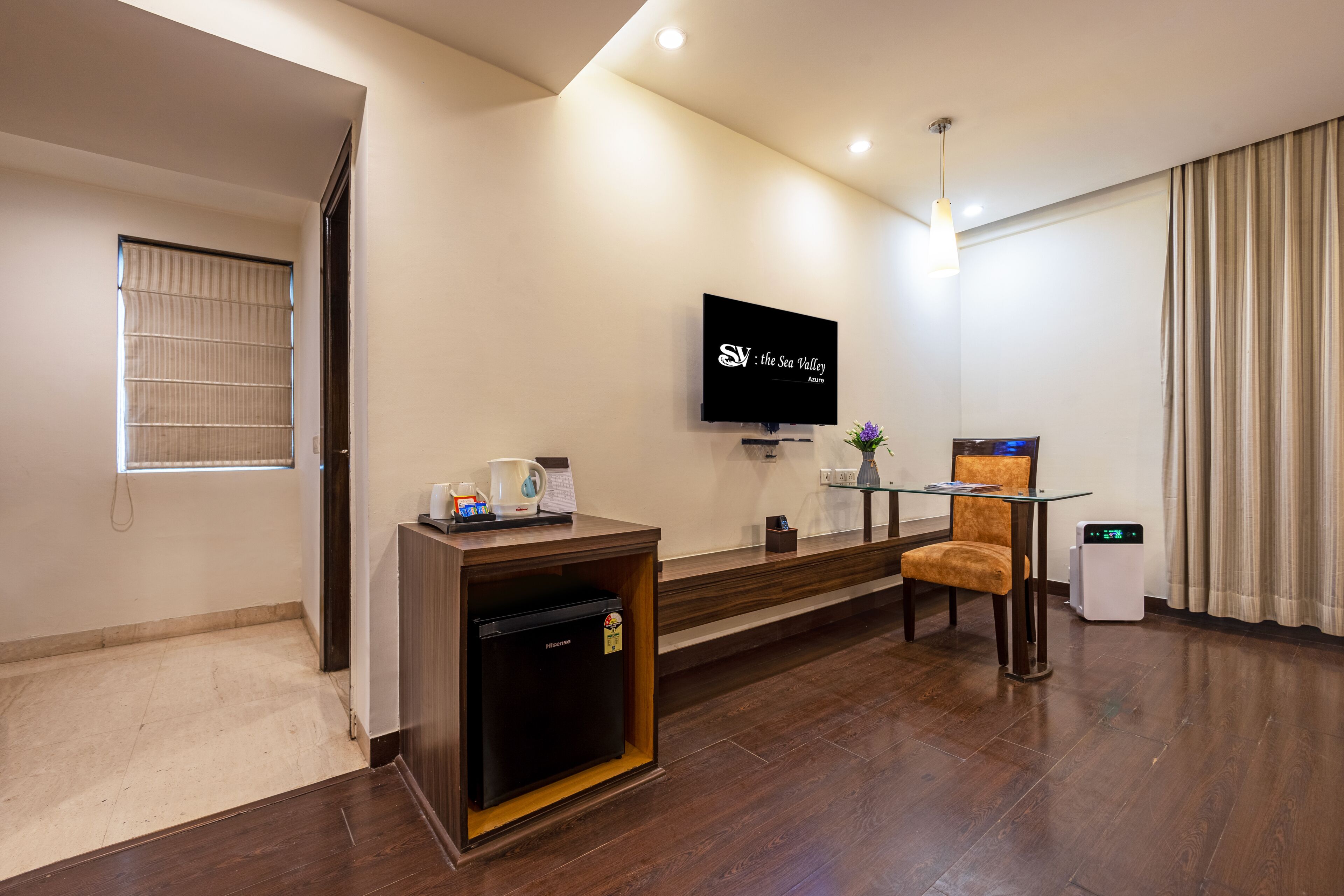 A hotel room with a flat-screen TV mounted on the wall, a small refrigerator, a coffee station, a glass table with a chair, and a window with blinds.