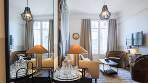 Ein modernes Wohnzimmer mit Sofa, Sesseln, Couchtisch und Fernseher. Zwei Lampen, ein dekorativer Spiegel und eine Kristallleuchter sind vorhanden.