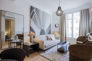Een moderne woonkamer met een bank, salontafel en stoelen. Er is een grote spiegel, een kroonluchter en een uitzicht op buiten door grote ramen.