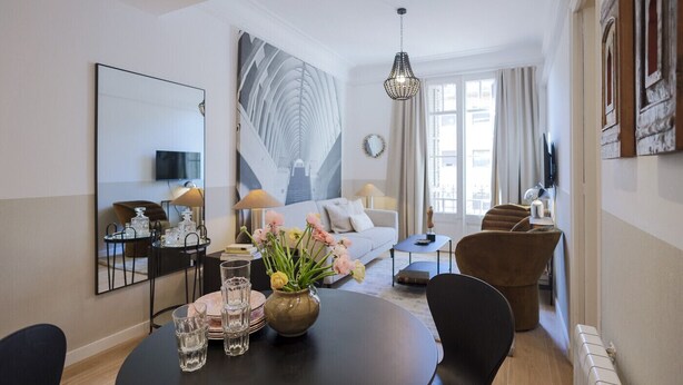 Ein modernes Wohnzimmer mit einer Couch, einem Esstisch und Stühlen. An der Wand hängt ein großes Gemälde, darüber ein Kronleuchter und ein Wandfernseher.