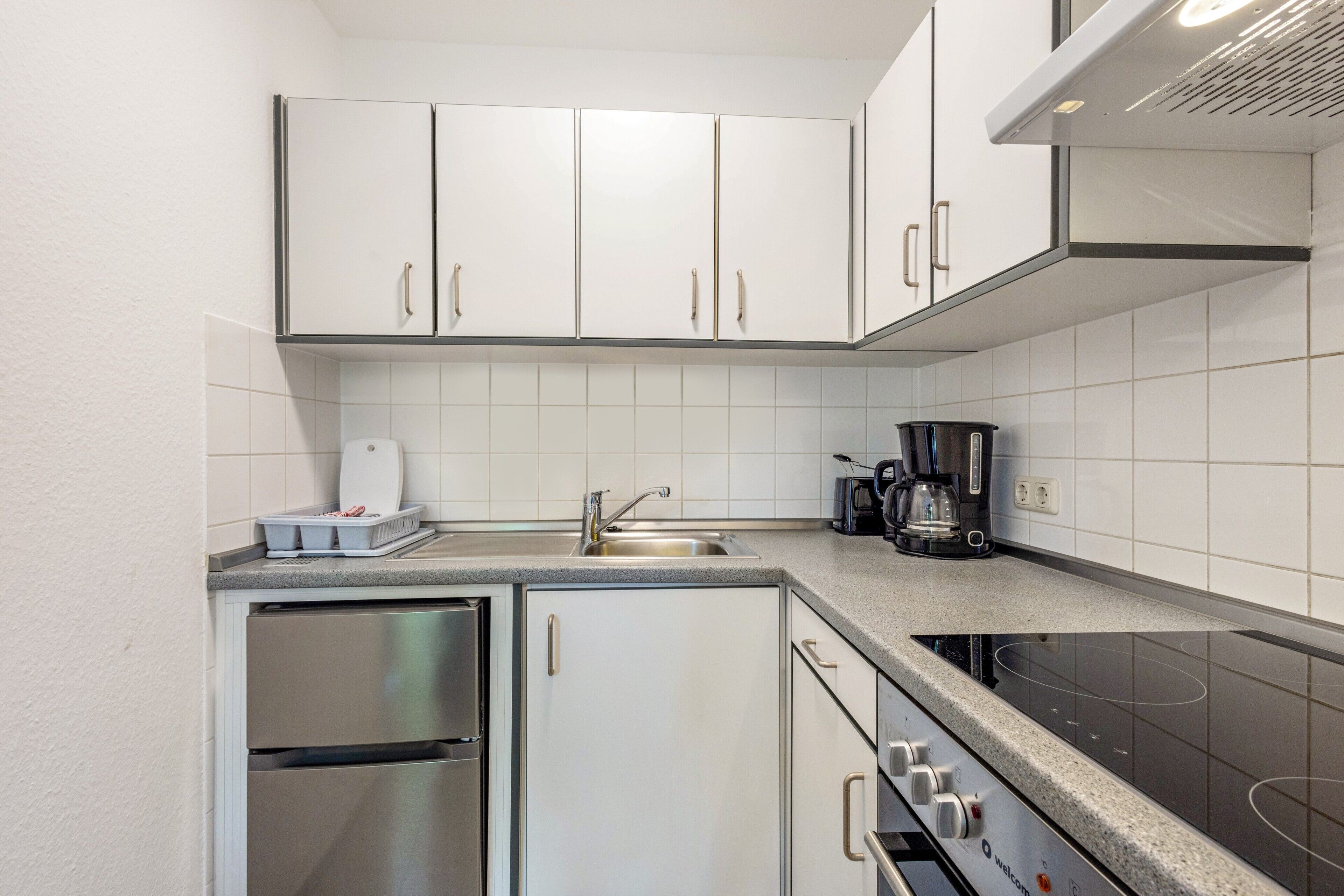 Una cuina compacta amb armaris blancs, un refrigerador d'acer inoxidable, un forn encastrat i una cafetera.