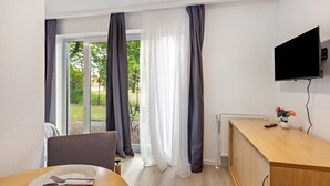 Huoneessa on ruokapöytä, tuoleja, seinälle asennettu televisio, kaappi ja ikkuna, jossa on verhot.