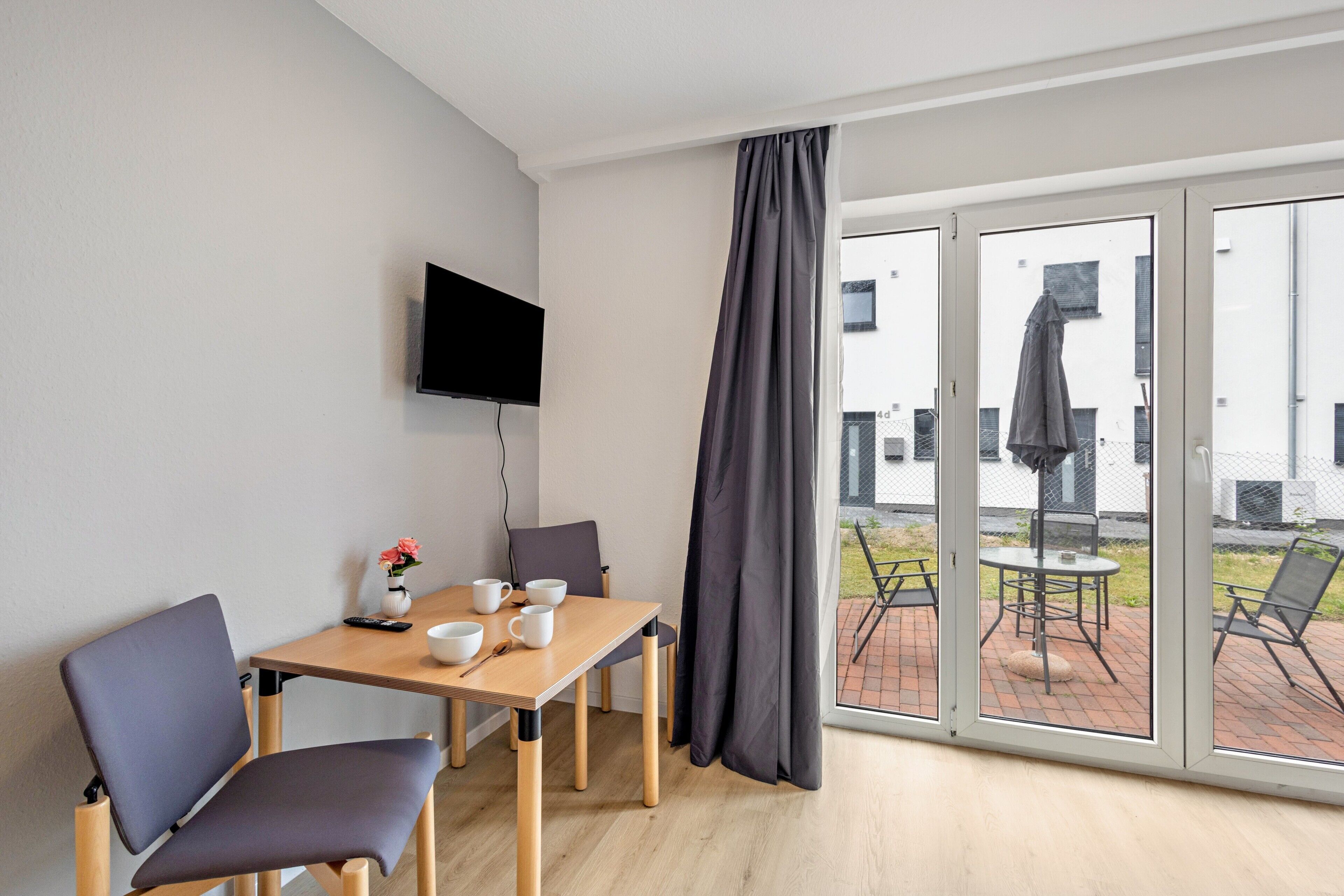 Een moderne kamer met een houten tafel, twee stoelen, een flatscreen televisie en een glazen schuifdeur die leidt naar een buitenruimte.