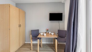 Una habitación con un armario de madera, una mesa con sillas, un televisor de pantalla plana montado en la pared y un ventanal con cortinas.