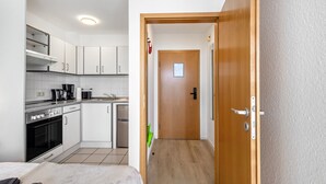 Una cocina compacta con armarios blancos, una estufa y un lavabo. Una puerta de madera da a un pasillo con un interruptor de luz y un ventanal pequeño.