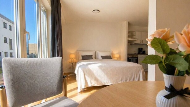 Una habitación de hotel con cama, mesitas de noche, una silla, una mesa y un florero con flores.