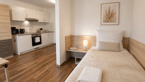 Una habitación de hotel moderna con suelo de madera, una cama con edredón blanco, una mesita de noche con lámpara y una zona de cocina al fondo.