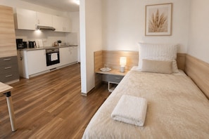 Ein modernes Hotelzimmer mit Holzboden, einem Bett mit weißer Bettdecke, einem Nachttisch mit Lampe und einer Küchenzeile im Hintergrund.