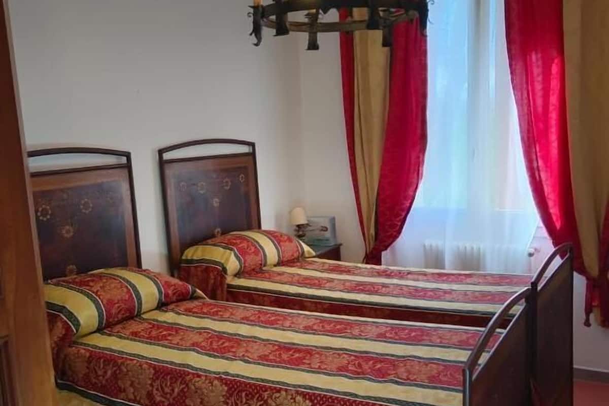 A room with two beds, a chandelier, red curtains, and a patterned rug.
