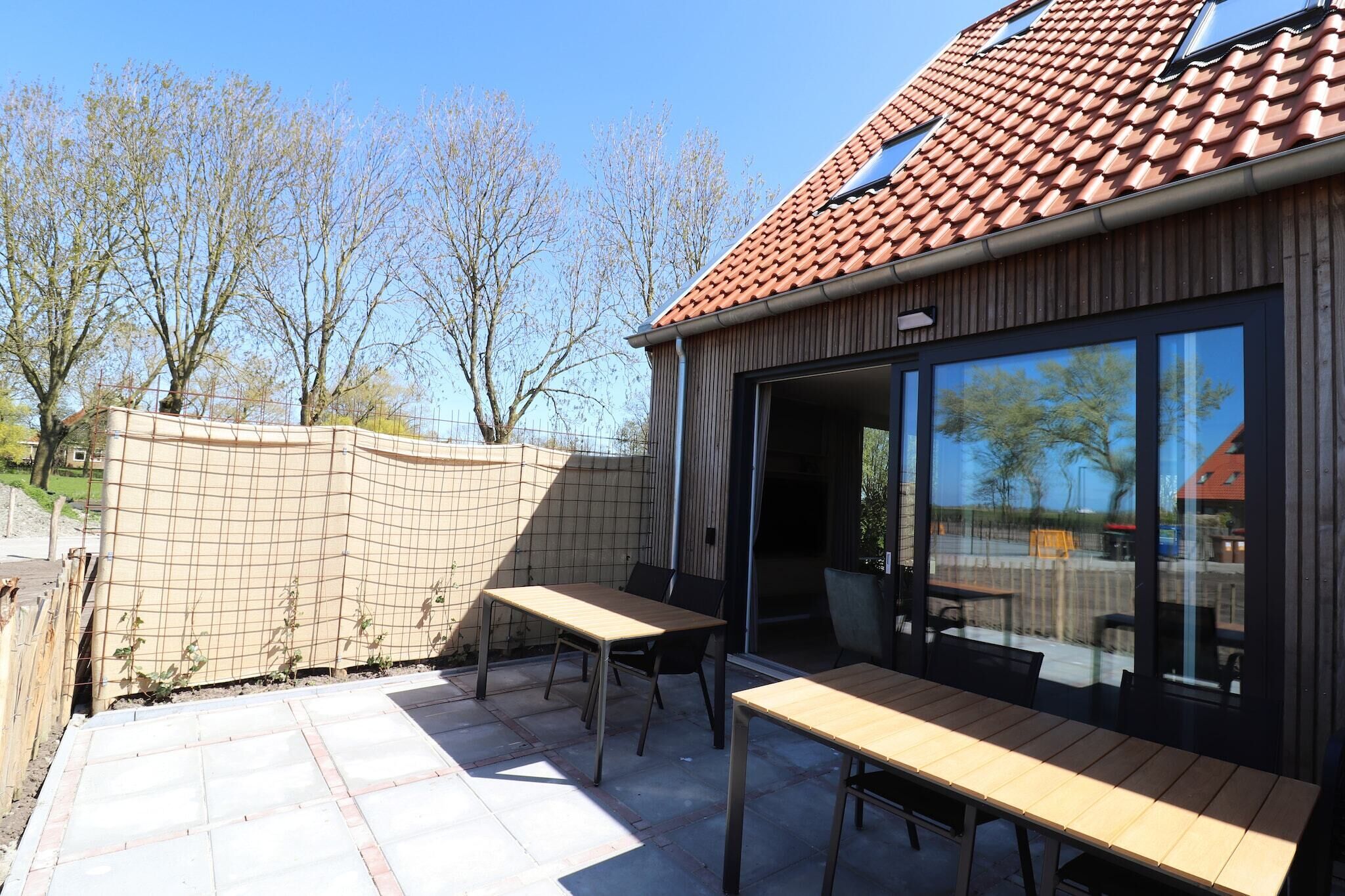 An outdoor patio with wooden tables and chairs, a brick wall, and a large glass door leading to a covered area.