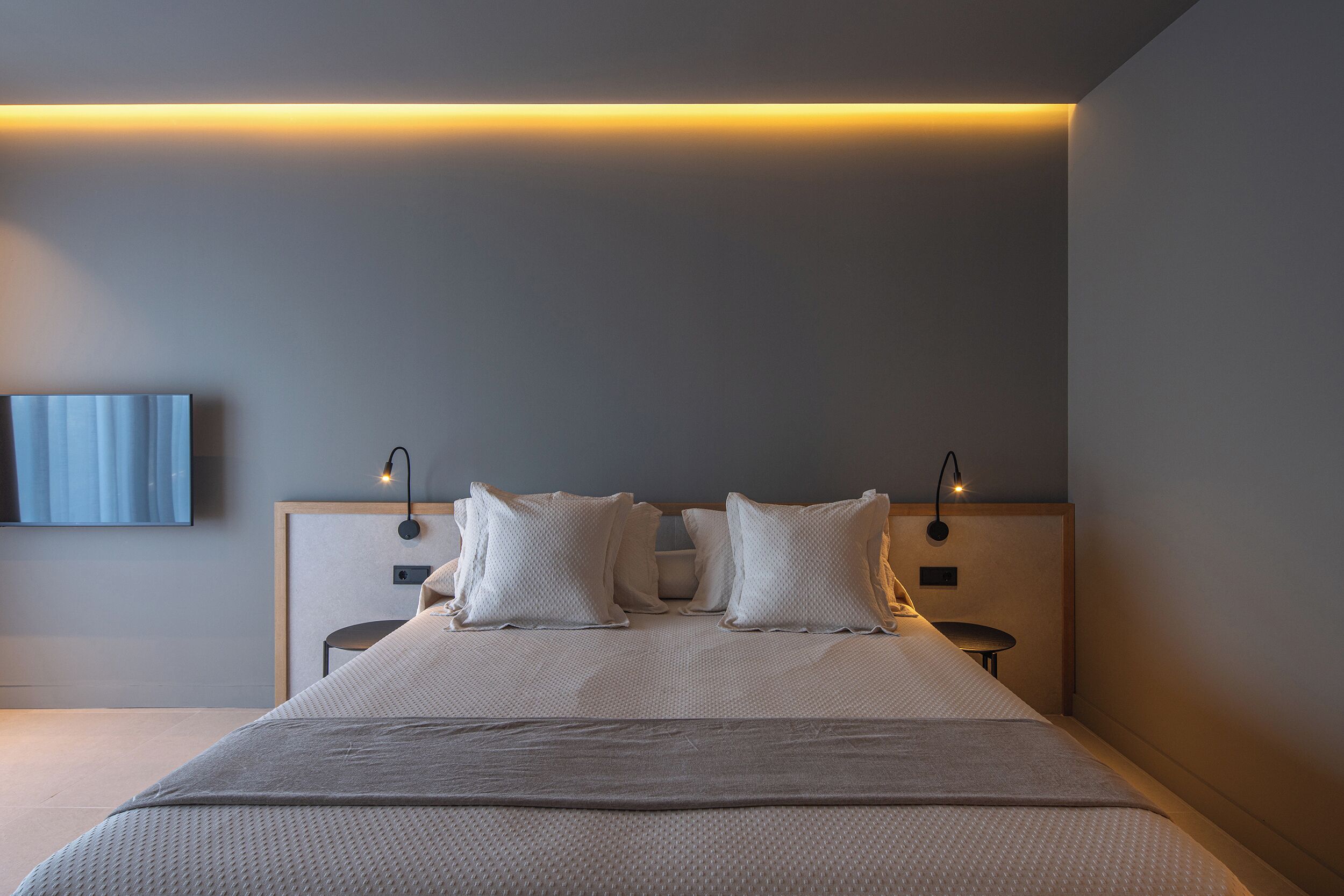A modern bedroom with a large bed, two bedside tables, a wall-mounted TV, and ambient lighting.